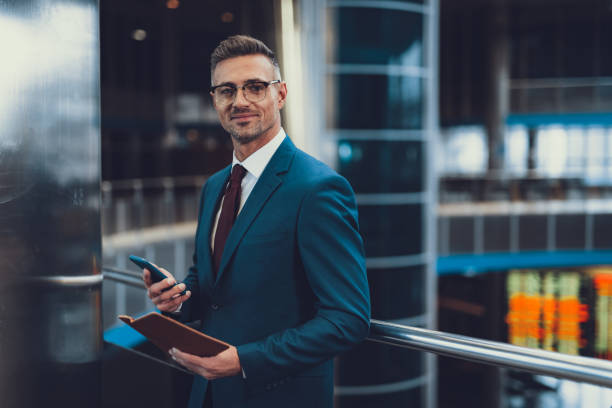 Attractive, adult man in chic formal wear standing inside business center. He looking at camera and holding telephone with document in hands