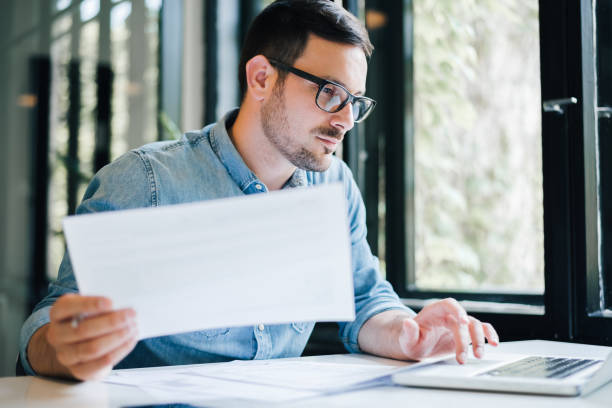 Serious pensive thoughtful focused young casual business accountant bookkeeper in office looking at and working with laptop and income tax return papers and documents